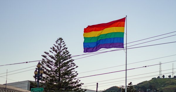 A large rainbow pride flag waving in an urban setting, representing LGBTQ+ community visibility and solidarity
