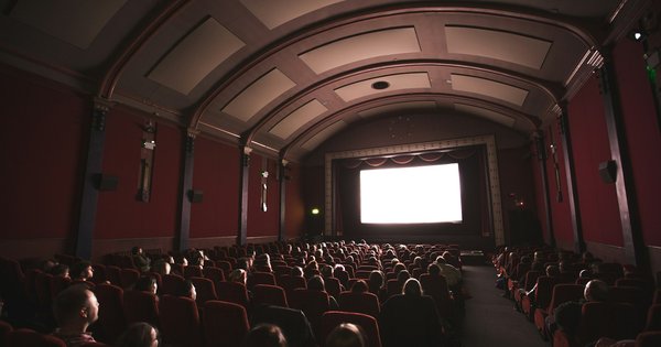 An audience seated in red velvet cinema seats, silhouetted against a bright screen in a darkened auditorium