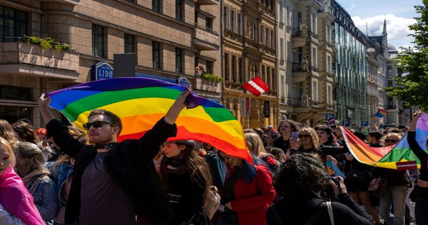 A dense crowd waving rainbow flags fills a broad avenue during a Pride march in Madrid under a bright summer sky
