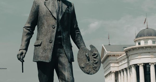 A bronze statue along the Vardar River in central Skopje, North Macedonia, with neoclassical government architecture in the background.