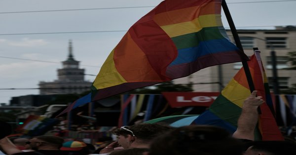 A rainbow Pride flag held aloft in front of a government building under a bright sky.