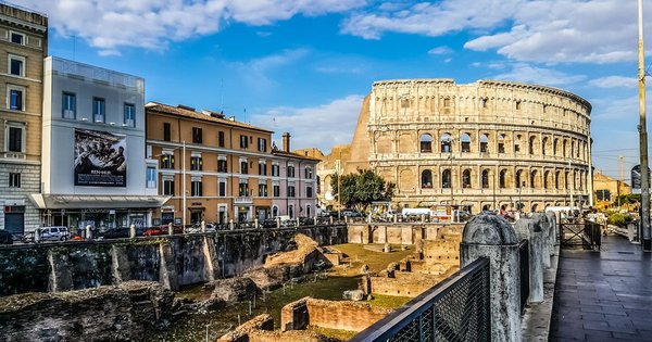 A huge crowd marching with rainbow flags past historic Roman architecture under bright sun.