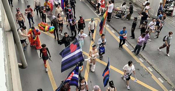 Pride marchers carrying a large rainbow flag through a sunlit street in Thailand