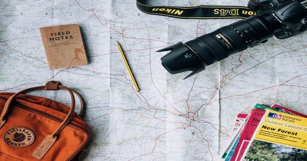 Travel flatlay with camera, field notes, maps, and a backpack spread on a road map